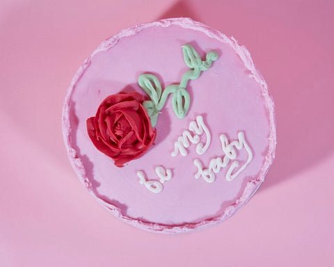 A single-tier, light pink cake with a textured edge and a romantic design. It features a red buttercream rose with green leaves and stem on the left side. The words "be my baby" are written in white icing in a playful, cursive style, creating a sweet and endearing message. The cake sits against a matching pink background, enhancing its soft, affectionate theme.