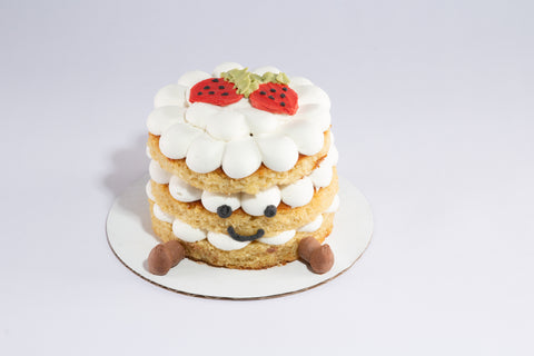 A whimsical three-tier vanilla cake with scalloped white frosting, decorated with red strawberry designs on top, chocolate feet at the base, and a cute smiley face. The cake is displayed on a white round board against a light gray background.