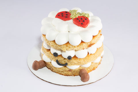 Front view of a cheerful three-tier strawberry shortcake with petal-like white frosting, two red strawberries with black seeds and green leaves on top, chocolate feet, and a playful smiley face made with black icing. Inspired by the Jellycat Amuseable Birthday Cake plush toy.