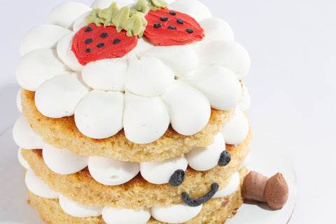 Close-up angle of a three-tier cake with fluffy white frosting, decorated with two red fondant strawberries with black seed details and green leaves. The cake features a smiling face with black icing eyes and a smile, sitting on a white cake board.