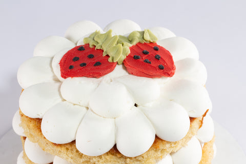Detail view of the top tier of a whimsical strawberry cake showing the red fondant strawberries with black seed details and green leaf accents nestled on white scalloped frosting. The golden vanilla cake base is visible beneath the white frosting.