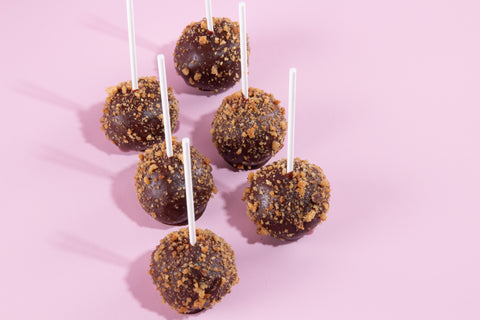 Six cake pops dipped in dark chocolate coating and rolled in golden-brown crushed cookie or graham cracker crumbs, displayed on white sticks against a light pink backdrop.