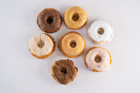 A top-down view of seven donuts arranged in a circular pattern. Each donut features a different topping, such as chocolate drizzle, cinnamon sugar, powdered sugar, and pink icing with sprinkles.