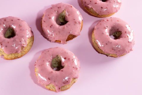 Four pink raspberry-glazed lemon donuts arranged on a light pink background. The donuts feature a golden cake base with visible poppy seeds and a glossy pink raspberry glaze with small red fruit pieces.