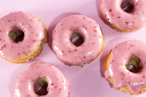 Close-up view of five raspberry-glazed lemon donuts on a pink surface. The donuts show a light golden cake texture with dark poppy seed specks and are topped with a shiny pink raspberry icing.