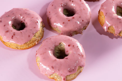 Four pink raspberry-glazed lemon donuts displayed on a pink backdrop. The donuts' cake texture is visible through the holes, showing a moist lemon poppy seed base covered with smooth pink raspberry glaze.