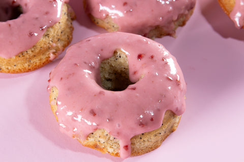 Close detail of a raspberry-glazed lemon donut with visible poppy seeds in the cake base. The pink glaze has small red fruit pieces and drips slightly down the sides of the golden donut.