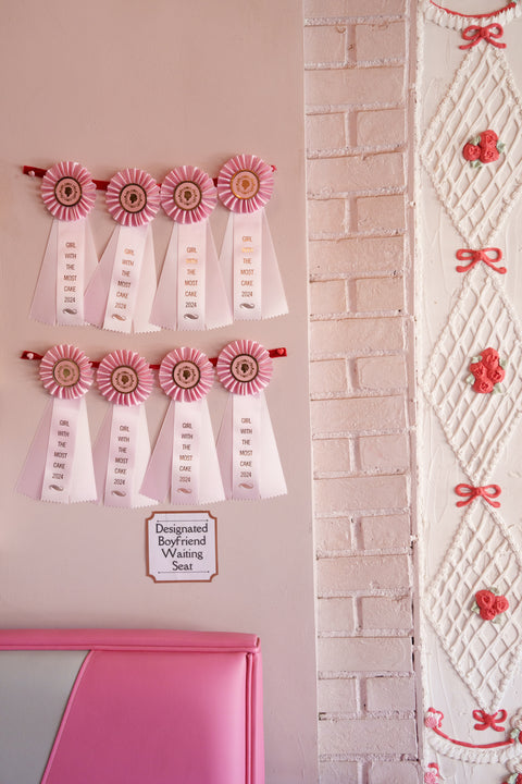 A wall adorned with several pink and white ribbons that say "Girl with the Most Cake 2024." Beneath the ribbons is a sign that reads "Designated Boyfriend Waiting Seat," and part of the wall is textured with a lace-like design featuring pink bows and flowers.