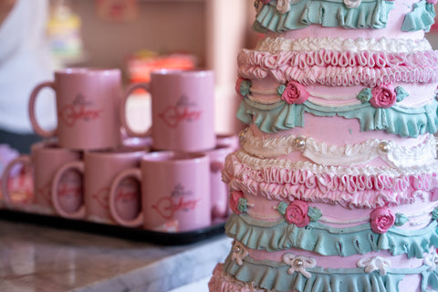 A close-up of a pink cake adorned with detailed pastel-colored frosting, decorated with ruffles, roses, and pearls. In the background, there are stacked pink mugs with a bakery logo.