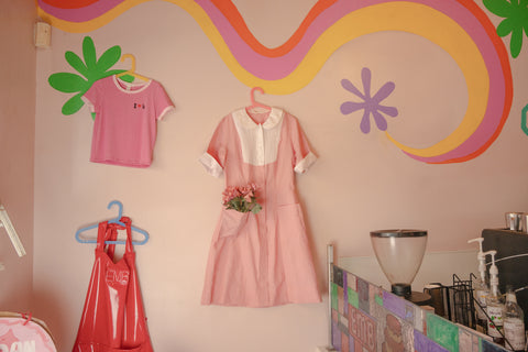 A decorative display with a pink uniform dress and an apron hanging on a wall. The wall is painted with colorful swirls and flower shapes. The dress has a white collar and is paired with a pink flower bouquet tucked into its pocket.