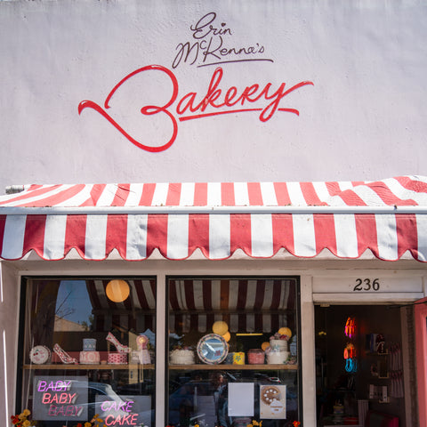 A view of the outside of Erin Mckenna's Bakery. There is a striped awning with a view of decadent cakes and decorations inside.