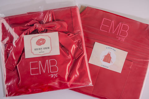 A vibrant red apron featuring the embroidered letters "EMB NYC" neatly packaged in clear plastic. The apron is showcased alongside a label featuring a red lip print and the text "Red Hot Apron by Erin McKenna." Both aprons are visibly folded, and one has a matching red ribbon tied in a bow for added style.
