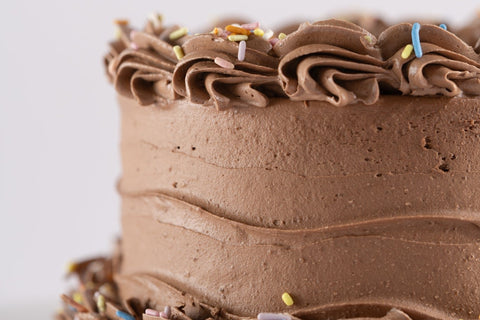 A close-up of a vegan and gluten-free chocolate-frosted cake with smooth sides and decorative piped chocolate swirls, topped with pastel-colored sprinkles.