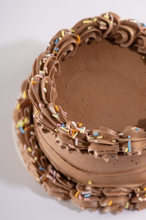 A top-down view of a vegan, gluten-free chocolate cake featuring piped chocolate buttercream swirls around the edge, decorated with colorful sprinkles.