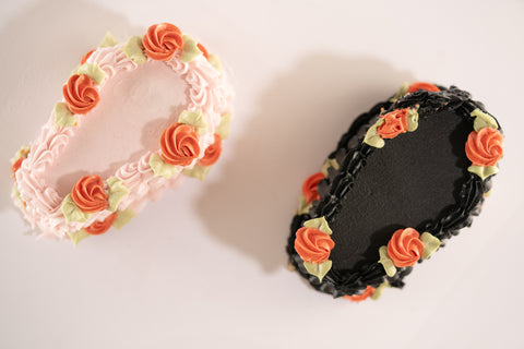 Two coffin-shaped Halloween mini cakes, one pink and one black, both decorated with orange roses and green leaves, perfect for a spooky celebration.