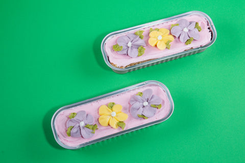 Two rectangular mini cakes with pink frosting topped with decorative purple and yellow flower blossoms and green leaves on a bright green background.