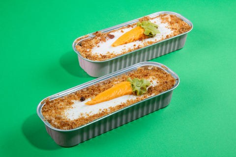 Mini cakes in aluminum tins displayed on green background, featuring carrot cakes with decorative orange carrots and cream frosting with cookie crumb borders.