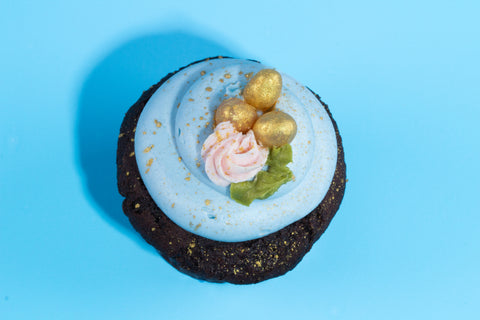 Chocolate cupcake with light blue frosting topped with decorative golden eggs, a small pink rosette, and green leaf accent against a bright blue background.