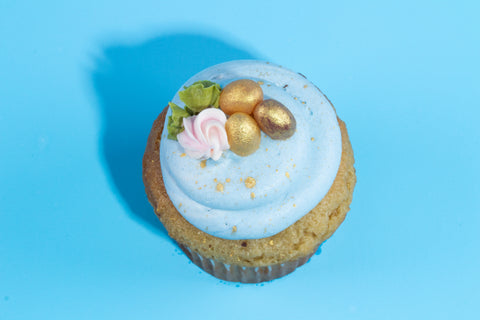 Vanilla cupcake with light blue frosting topped with golden eggs, pink piping, and green leaf accent against a sky blue background.