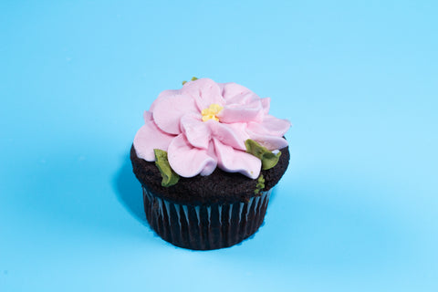 Chocolate cupcake decorated with an intricate pink flower blossom and green leaves against a bright blue background.