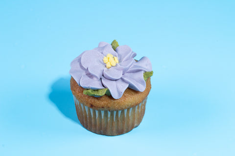 Vanilla cupcake with a detailed purple flower blossom decoration and yellow center against a bright blue background.