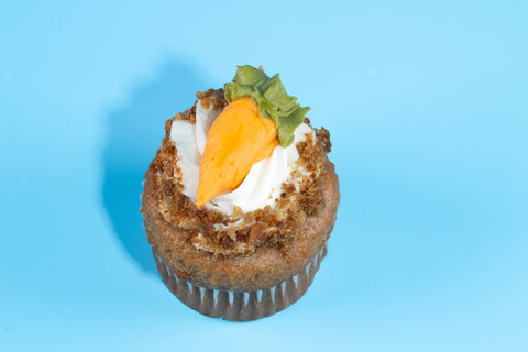 Close-up of a carrot cake cupcake with white frosting, cookie crumble topping, and PIPED orange carrot decoration against a blue background.
