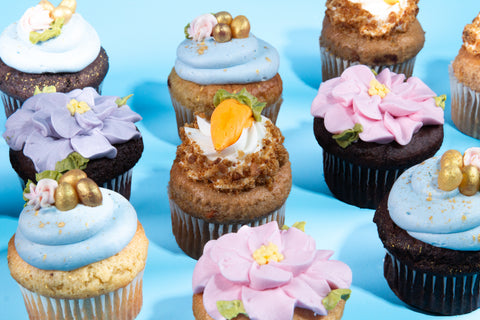 Close-up arrangement of various Easter cupcakes featuring purple and pink frosting flowers with yellow centers, blue frosted cupcakes with golden eggs, and carrot cake cupcakes on blue background.