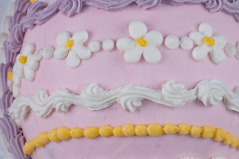 Close-up view of a pink egg-shaped cake decorated with white shell piping, yellow dot borders, lavender frosting accents, and a small yellow flower in the center.