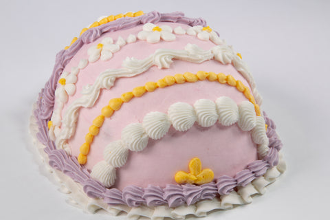 Angled view of a pink Easter egg cake featuring decorative piping in white, lavender, and yellow with small daisy flowers and alternating border designs.
