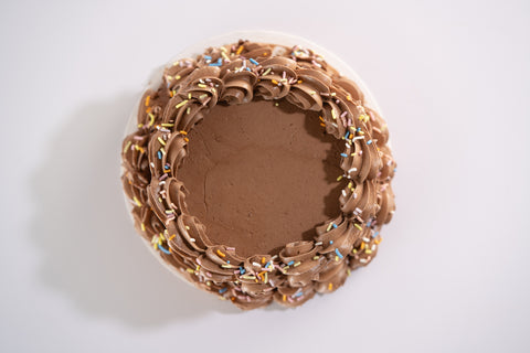 A top-down view of a vegan and gluten-free chocolate cake with piped chocolate buttercream swirls around the edges, sprinkled with colorful confetti.