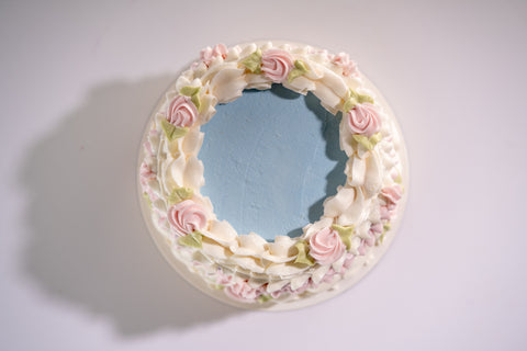 A top-down view of a vegan and gluten-free cake with pastel blue frosting, surrounded by white and pink piped buttercream swirls and floral decorations.
