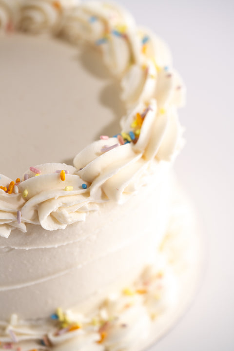 A close-up of a vegan and gluten-free white frosted cake with piped swirls around the edges, accented with colorful sprinkles.