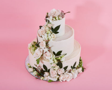 A three-tiered white cake adorned with delicate pink and white roses cascading down the layers, accented with lush green leaves and small sprigs of filler flowers. The background is a soft pink, giving the entire setup a romantic and elegant look.