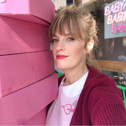 A woman with light brown hair styled in a loose bun and bangs poses next to a tall stack of pink boxes. She is wearing a white shirt with a red sweater and light red lipstick. In the background, there is a neon sign that says "Baby Baby Baby."