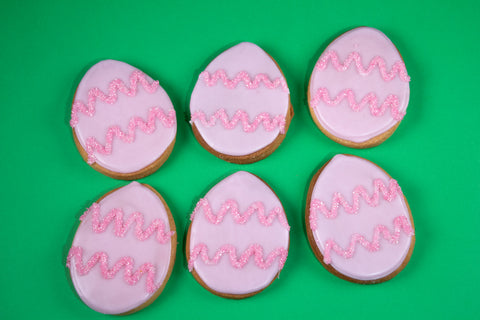 Collection of six oval egg-shaped cookies with glossy pale pink icing adorned with sparkling hot pink sugar zigzags, arranged in a playful pattern against a vibrant emerald green background.