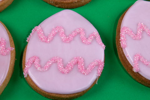 Collection of six oval egg-shaped cookies with glossy pale pink icing adorned with sparkling hot pink sugar zigzags, arranged in a playful pattern against a vibrant emerald green background.