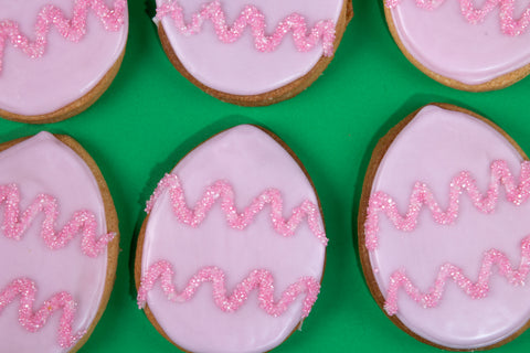 Collection of six oval egg-shaped cookies with glossy pale pink icing adorned with sparkling hot pink sugar zigzags, arranged in a playful pattern against a vibrant emerald green background.