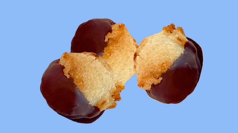 Three chocolate-dipped macaroons are displayed against a blue background. Each macaroon is half-covered in rich dark chocolate, with the golden coconut texture visible on the uncovered portions, highlighting Erin McKenna's Bakery's gluten-free, kosher, and vegan Passover offerings.