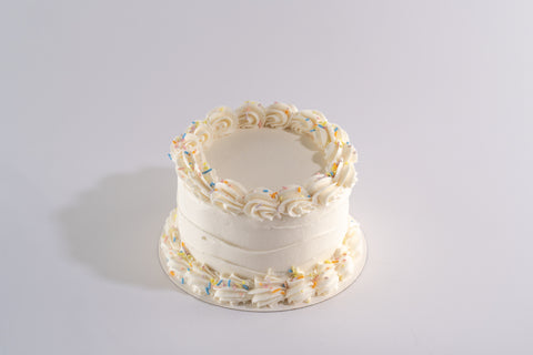 A vegan and gluten-free white frosted cake with smooth buttercream, decorated with piped white swirls on the top and bottom edges, and colorful sprinkles.