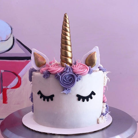 A unicorn-themed cake with a smooth white frosting base, featuring closed black eyelashes, a swirl of pink and purple buttercream rosettes as a mane, and a golden spiral horn at the center. The cake is topped with unicorn ears that have a golden accent on the inside, creating a whimsical and magical appearance.