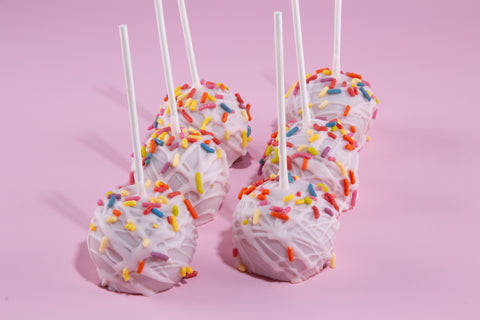 Six birthday cake-style cake pops with white coating generously covered in multicolored sprinkles, mounted on white sticks and displayed on a pastel pink surface.