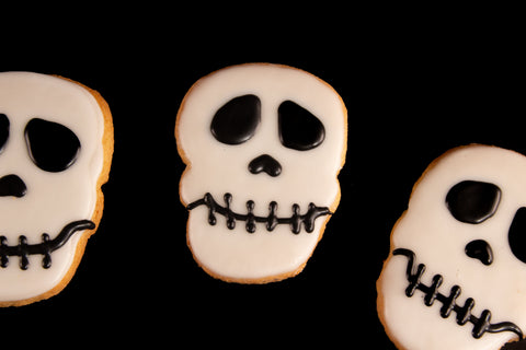 Gluten-free vegan skull-shaped sugar cookies with white frosting and black icing skeleton faces on black background - allergy-friendly Halloween skeleton cookies