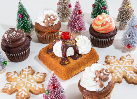 Festive holiday dessert display with gluten-free cupcakes, gingerbread cookies, and cake arranged among miniature Christmas trees in green, pink, silver, and gold with tiny ornaments creating a winter wonderland scene.
