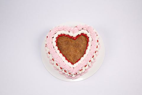 Heart-shaped chocolate chip cookie cake decorated with pink and white vegan buttercream rosettes and red piping details on a white cake board