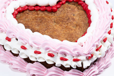 Heart-shaped chocolate chip cookie cake decorated with pink and white vegan buttercream rosettes and red piping details on a white cake board