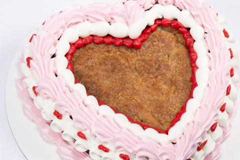 Heart-shaped chocolate chip cookie cake decorated with pink and white vegan buttercream rosettes and red piping details on a white cake board