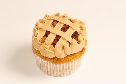 Close-up of a single vegan apple pie cupcake featuring a lattice-design vanilla frosting top, decorative piped edges, and visible spiced apple filling through the lattice pattern on a light background