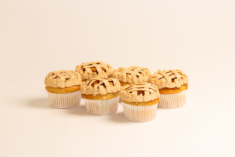Six vegan gluten-free apple pie cupcakes arranged in a row, each decorated with lattice-pattern frosting and piped borders, displayed on a neutral cream background