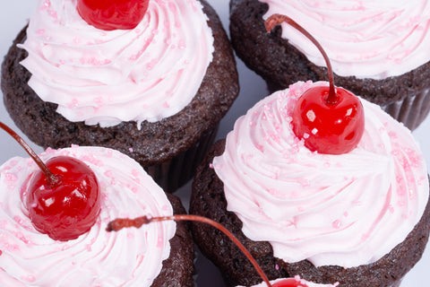 Chocolate cupcakes topped with swirled pink dairy-free buttercream frosting and glossy maraschino cherries on white background