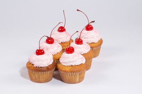 Vanilla cupcakes topped with swirled pink dairy-free buttercream frosting and glossy maraschino cherries on white background